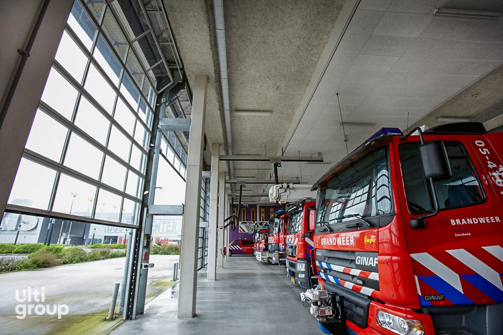 Red fire trucks parked inside a station with large glazed sectional doors.