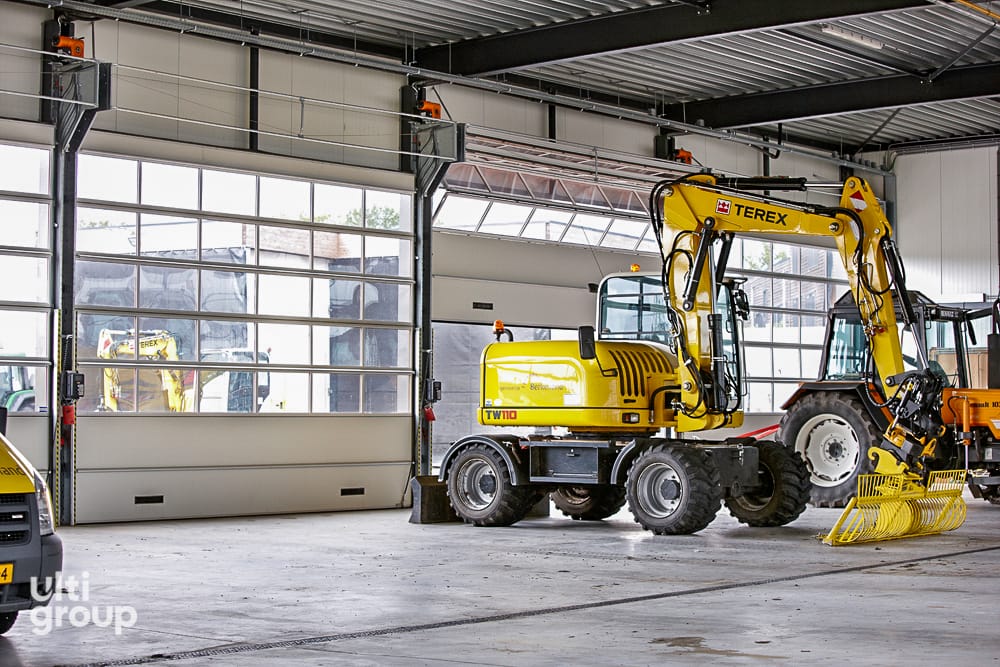 Yellow Terex excavator parked inside a workshop with large sectional glass doors.