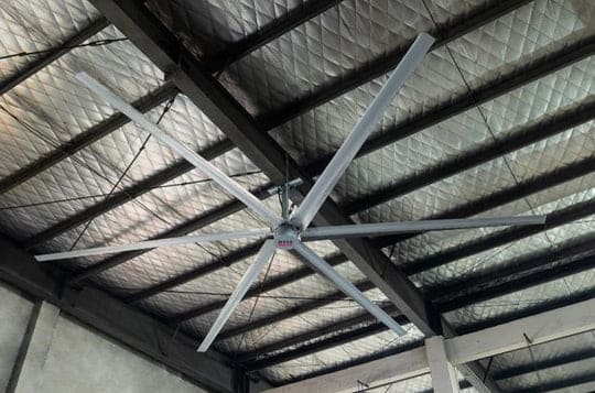 Large industrial ceiling fan with six silver blades mounted to a warehouse roof.