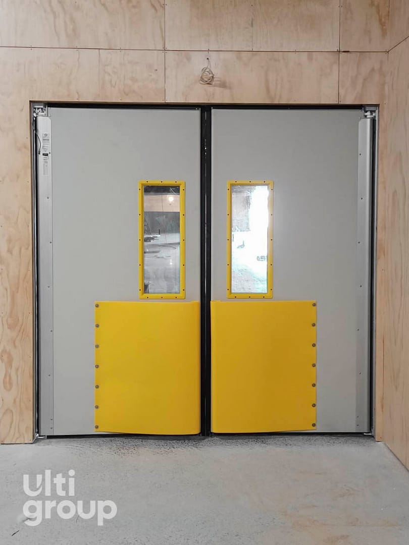 Grey double-leaf impact doors with yellow kick plates and vision panels.