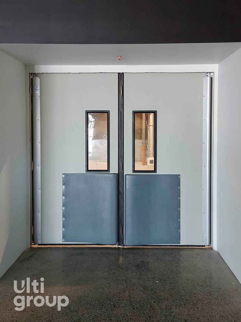 Grey double-leaf impact traffic doors with vision panels and kick plates.
