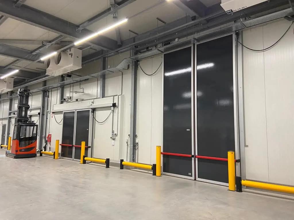 Large grey insulated bi-parting doors in a warehouse with yellow safety bollards.