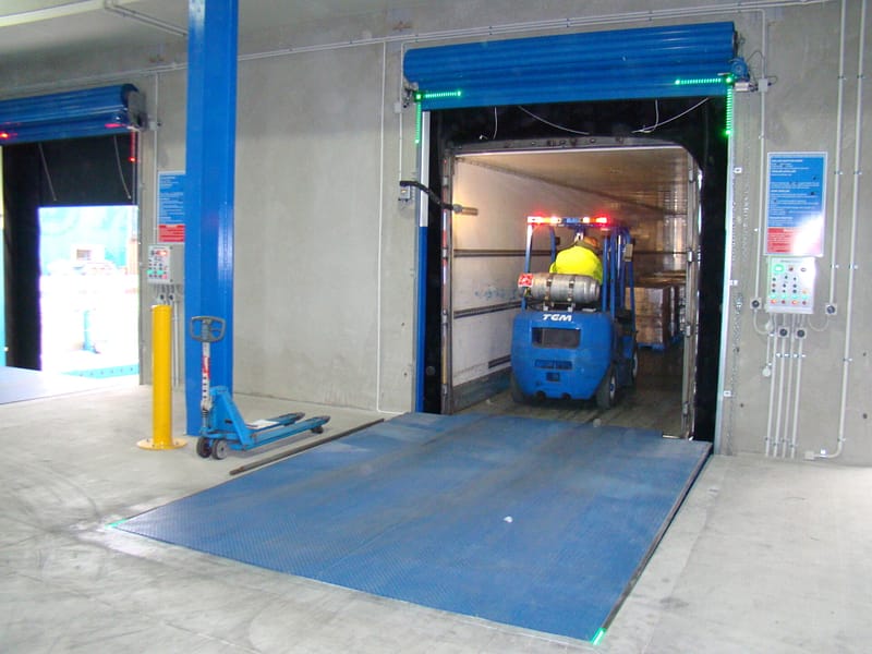 Blue forklift entering a truck trailer through a loading dock with a blue dock leveller.