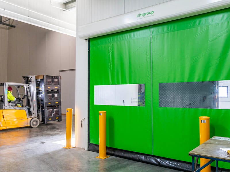 Green high-speed industrial door with vision panels and yellow safety bollards.