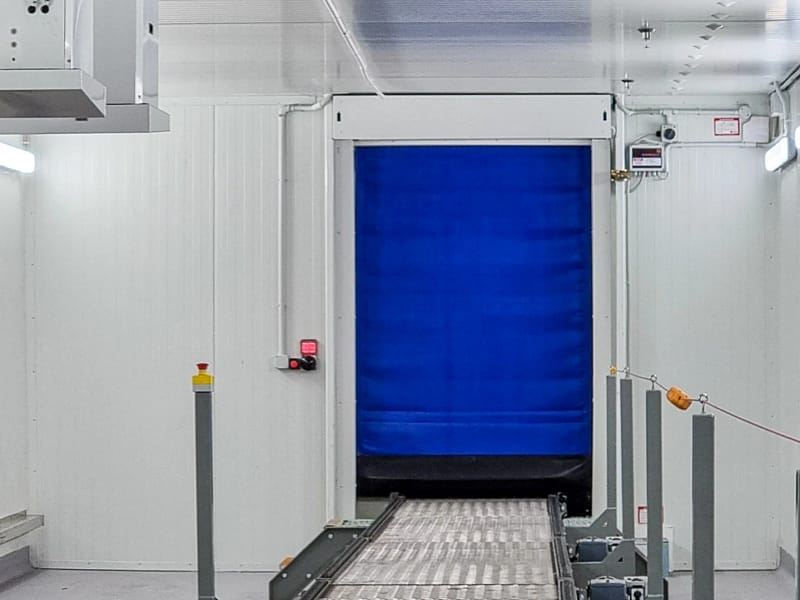 Blue high-speed roll door installed above a metal conveyor belt in a cold storage facility.