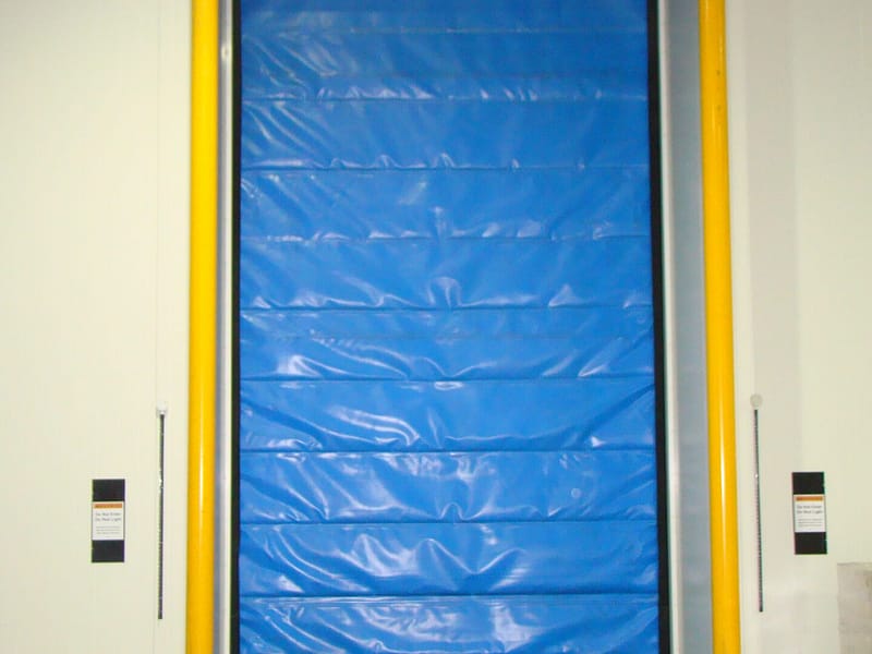 Blue FasTrax FR Door installed in a cold storage facility with yellow safety bollards.