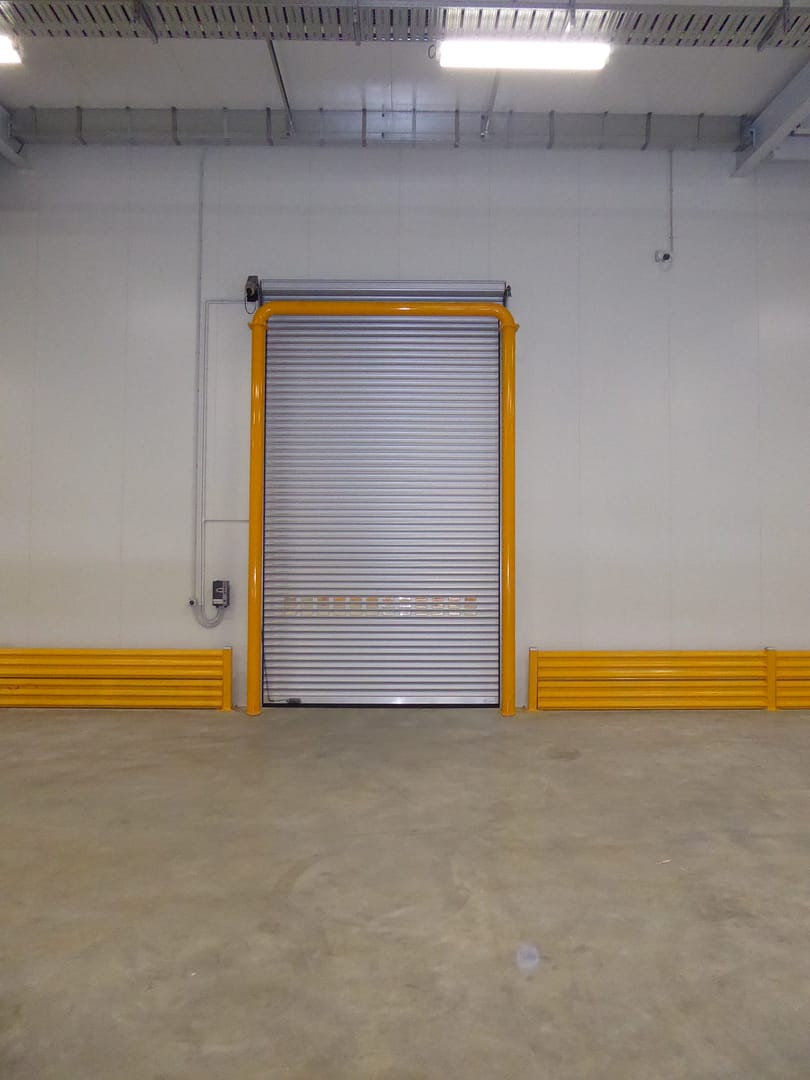 Insulated roller shutter door with yellow safety bollards and wall protection.