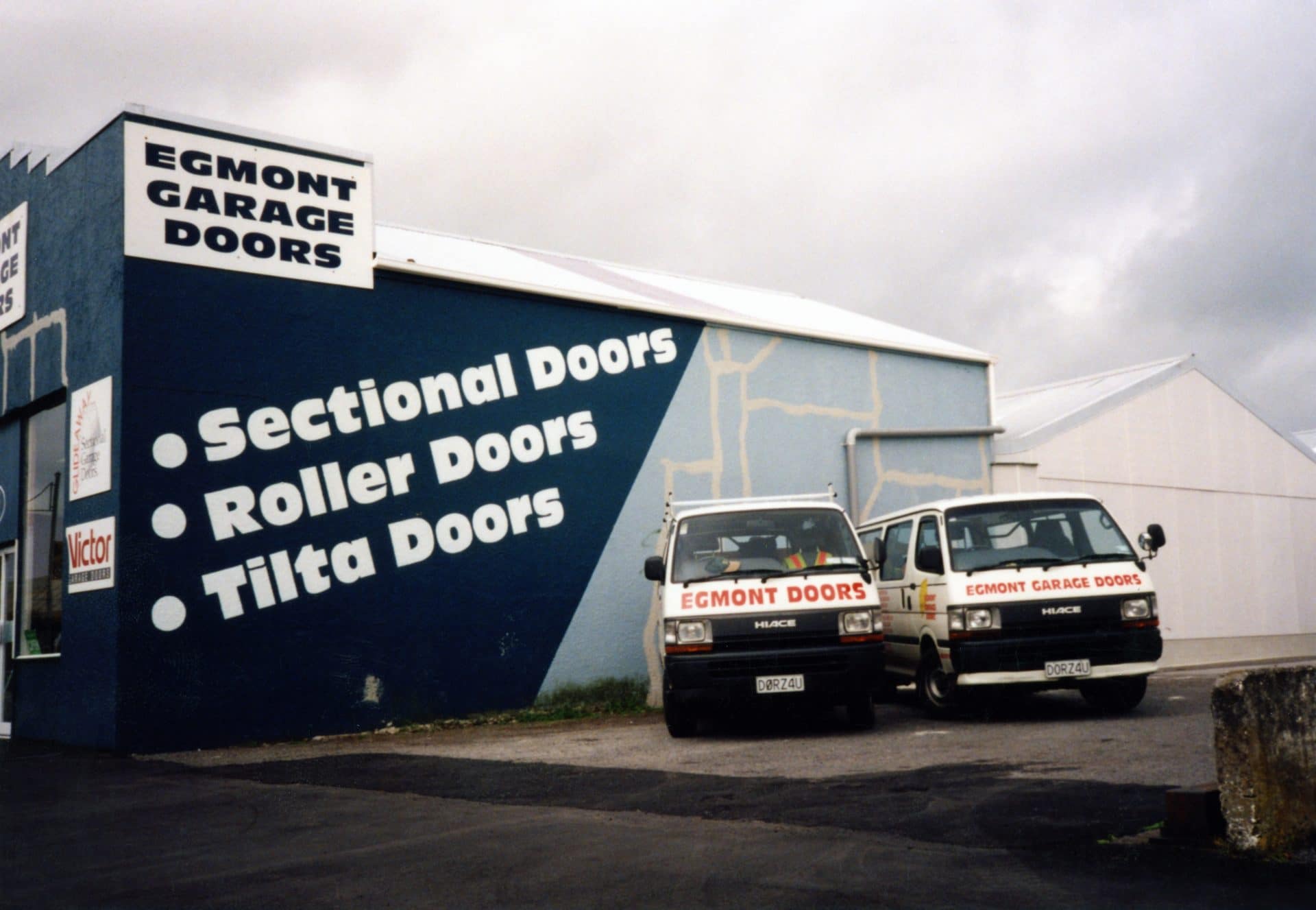 Two Egmont Garage Doors service vans parked outside a blue and white building.